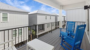 A covered balcony with a white table and blue chairs, overlooking a row of white buildings with windows. - The Hive a one-minute walk to the beach! Two community pools & nearby Lagoon access (Gulf Shores)