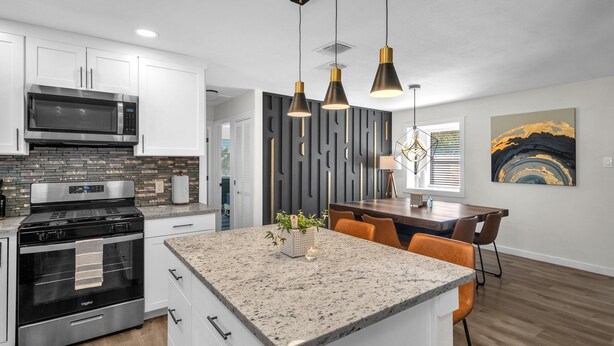 A modern kitchen with white cabinets, a granite countertop, a built-in stove, and a microwave. There is a dining area with a wooden table and chairs. - Lackland AFB Luxury - Sleeps 15 · Games · Big Yard (San Antonio)