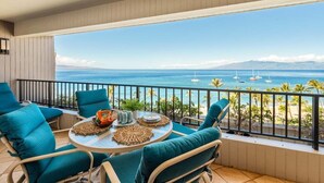 Un balcon avec vue sur l’océan et les voiliers, agrémenté de chaises turquoise et d’une table ronde où sont disposées des boissons et des en-cas.