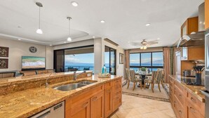 Une cuisine moderne dotée de plans de travail en granit, d’armoires en bois et d’un grand îlot central. Un coin repas offre une vue sur l’océan.