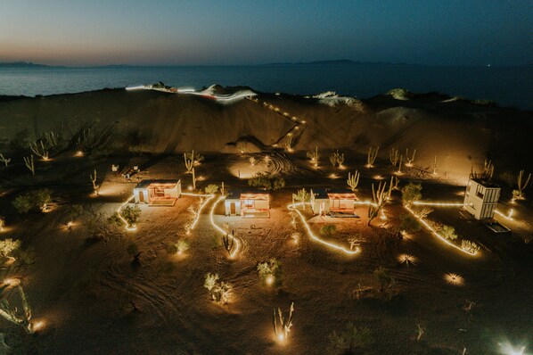 Experience the tranquil desert landscape with illuminated pathways connecting serene structures amidst the vast arid terrain and the distant ocean.