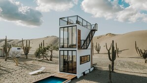 Una villa moderna de varios niveles en el desierto, con un salón envuelto en vidrio, piscina y una escalera que lleva a una terraza superior.