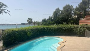 A swimming pool with a curved edge, surrounded by a hedge and a tiled patio area. - Apartment with air co and breathtaking sea view and direct beach access for 6 (SANARY SUR MER)