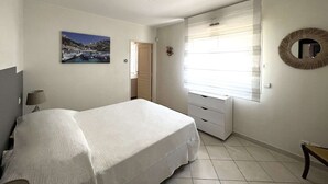 A bedroom with a bed, a bedside table, a dresser, a window with blinds, a wall-mounted mirror, and a picture of a coastal scene. - Apartment with air co and breathtaking sea view and direct beach access for 6 (SANARY SUR MER)