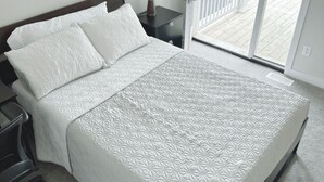 Una cama bien hecha con cubrecama blanco acolchado, una mesita de noche con una lámpara y vistas al balcón exterior a través de una puerta de cristal.