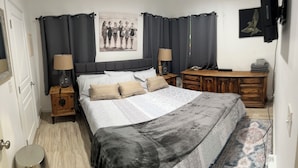 Quarto com cama, duas mesas de cabeceira, cômoda de madeira, quadro na parede e uma porta.