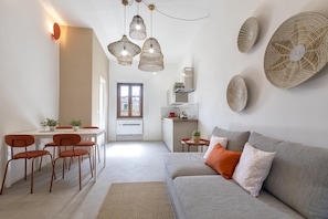 A modern living room with a grey sofa, orange cushions, and a dining area with a white table and red chairs. - Sardinia Re - Casa Pinuccia (Alghero)