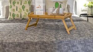 Un letto con una coperta grigia a trama decorativa, un vassoio di legno con una scatola etichettata “sardinia re” e due tazze bianche, e un cuscino con motivo cactus.