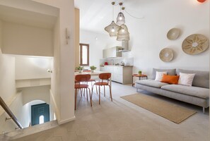 A modern living room with a sofa, chairs, and a dining area. There is a kitchen in the background with a window and a hanging light fixture. - Sardinia Re - Casa Pinuccia (Alghero)
