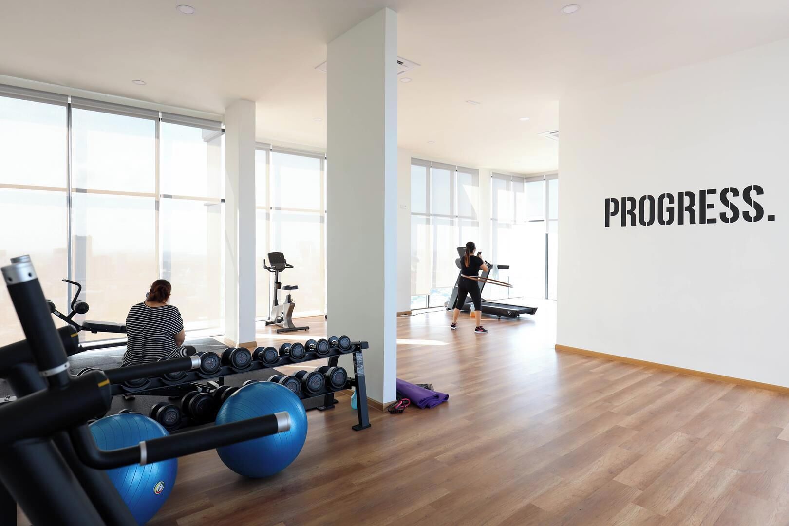 Un gimnasio con equipo de ejercicio, una mujer usando una cinta de correr y una pared con la palabra 'PROGRESS'.