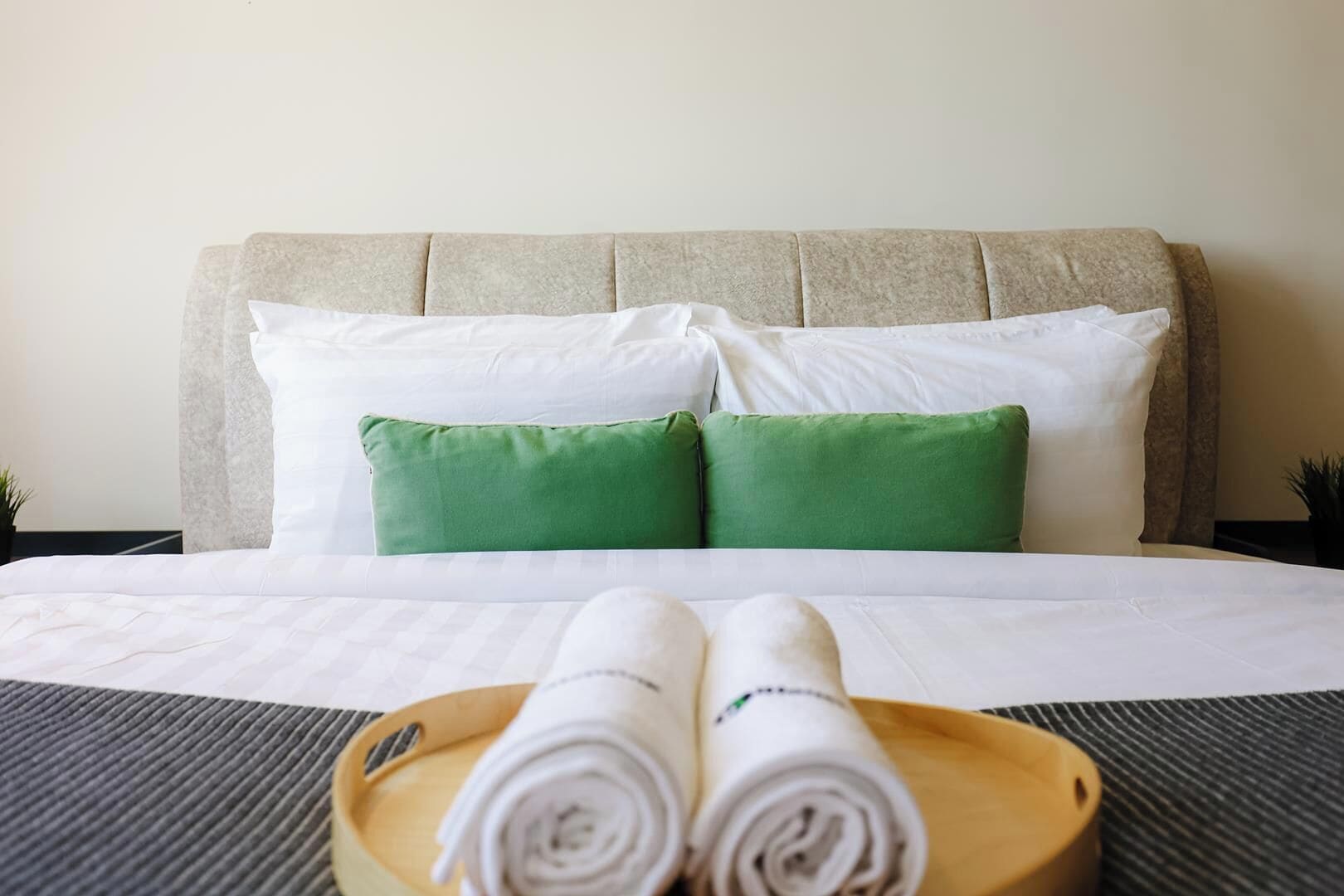 A neatly made bed with white and green pillows, a wooden tray with rolled towels, and a headboard.