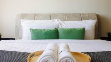 A neatly made bed with white and green pillows, a wooden tray with rolled towels, and a headboard.