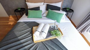 Un lit bien fait, recouvert d’une couverture grise, avec deux oreillers, un plateau contenant des serviettes et des verres, et une petite plante.