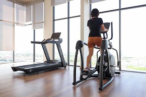 Une salle de sport intérieure spacieuse, équipée d’un tapis de course et d’un vélo elliptique, de grandes fenêtres et offrant une vue sur la ville.