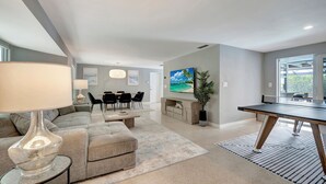 A modern living room with a sectional sofa, a dining area with a table and chairs, a large flat-screen TV, and a ping pong table.