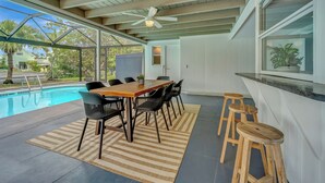 A covered outdoor dining area with a pool, wooden table, chairs, and stools.