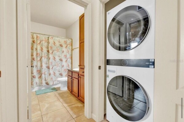 A laundry room with two stacked washing machines, a shower curtain with a scallop shell pattern, wooden cabinets, and a tiled floor.