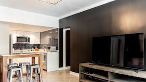 Cocina moderna con microondas, tostadora y lavamanos. Hay una zona de comedor con taburetes y un televisor en la sala de estar.