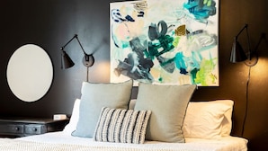 Un dormitorio moderno con un cuadro abstracto grande, dos lámparas de noche, un espejo redondo y una cama bien hecha con una colcha estampada.