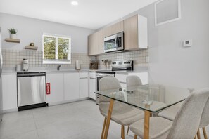 Une cuisine moderne avec des meubles blancs, un lave-vaisselle en acier inoxydable, un four à micro-ondes et un four traditionnel. On y trouve une table à manger en verre, accompagnée de deux chaises.