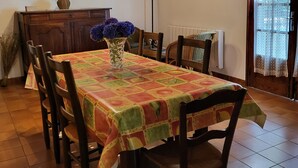 Un comedor con una mesa cubierta por un mantel colorido, rodeado de sillas de madera. Hay un jarrón con flores púrpuras sobre la mesa.