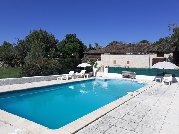 Une piscine rectangulaire bordée de carreaux, entourée d’une terrasse également carrelée de couleur blanche. On y trouve des chaises longues blanches et des parasols, et un bâtiment en pierre avec un toit carrelé en arrière-plan.