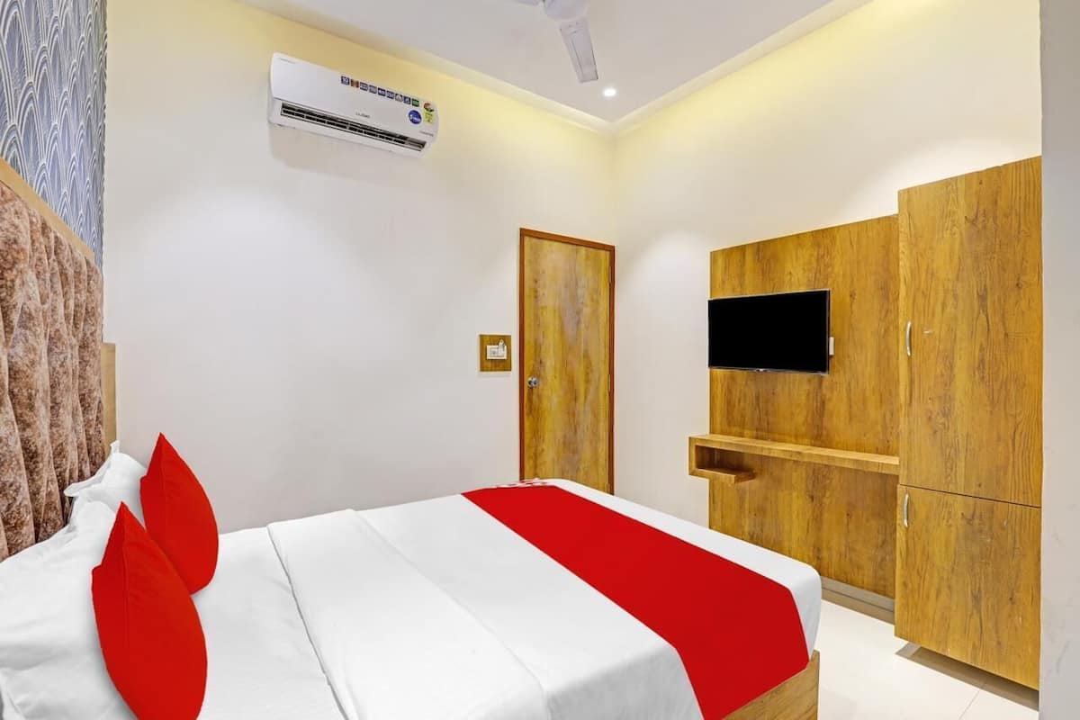 A hotel room with a bed, a TV mounted on a wooden cabinet, an air conditioning unit on the wall, and a door.