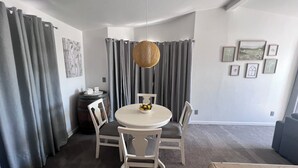 Et spiseområde med et rundt bord, fire stole, en hængende lampe, grå gardiner og billeder i rammer på væggen.