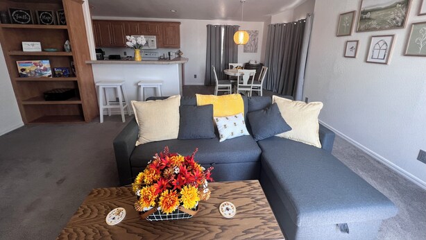 En moderne stue med en grå modulsofa, et træsofabord med blomsterarrangement, og et køkken i baggrunden.