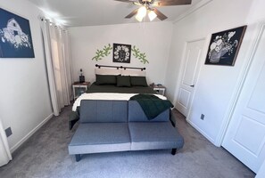 A bedroom with a bed, a grey sofa, a ceiling fan, and two nightstands.