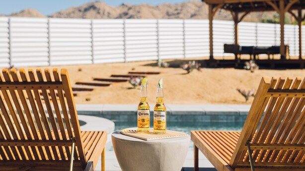 Un área junto a la piscina con sillones de madera y dos botellas de cerveza Corona sobre una mesa.