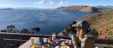 A table set with breakfast items and a person sitting at the table, overlooking a large body of water and hills.