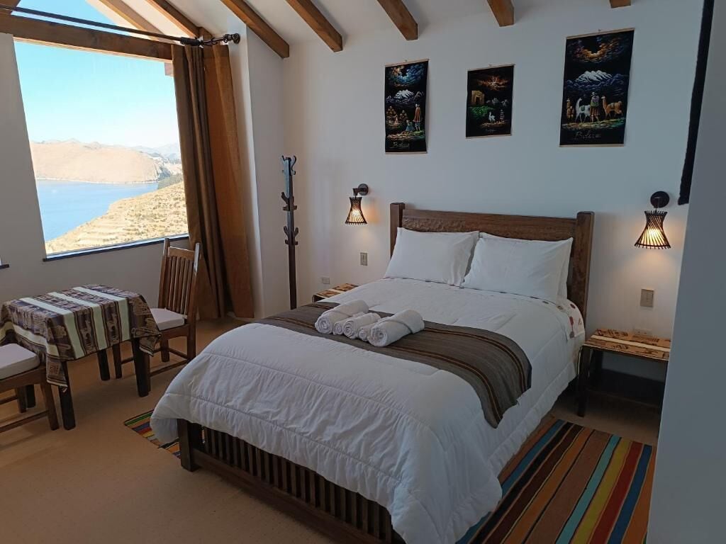 A bedroom with a large bed, two bedside tables, a chair, a small table, and a window with a view of the sea.