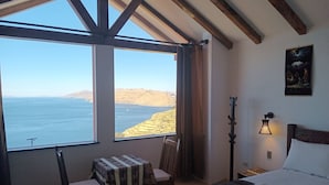 A bedroom with a large window overlooking a coastal landscape, a bed with white linens, a small table with a patterned tablecloth, and a wall-mounted lamp. - Nueva Luna - ISLA DEL SOL - Bolivia (Isla del Sol)