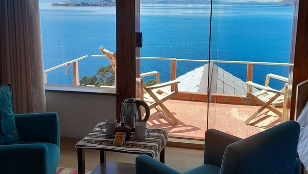 Quarto com uma grande janela com vista para a água e terra ao longe. Há uma mesa com chaleira e xícaras, duas cadeiras azuis e um tapete listrado no chão.