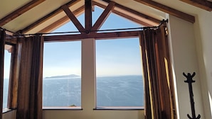 A room with a high ceiling and exposed wooden beams, large windows with curtains, and a view of a distant landscape. - Nueva Luna - ISLA DEL SOL - Bolivia (Isla del Sol)