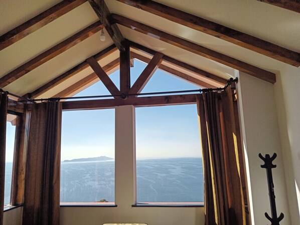 A room with a high ceiling and exposed wooden beams, large windows with curtains, and a view of a distant landscape. - Nueva Luna - ISLA DEL SOL - Bolivia (Isla del Sol)