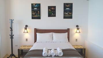 Una cama bien hecha con sábanas blancas y toallas enrolladas, cabecera de madera y dos mesitas de noche con lámparas.