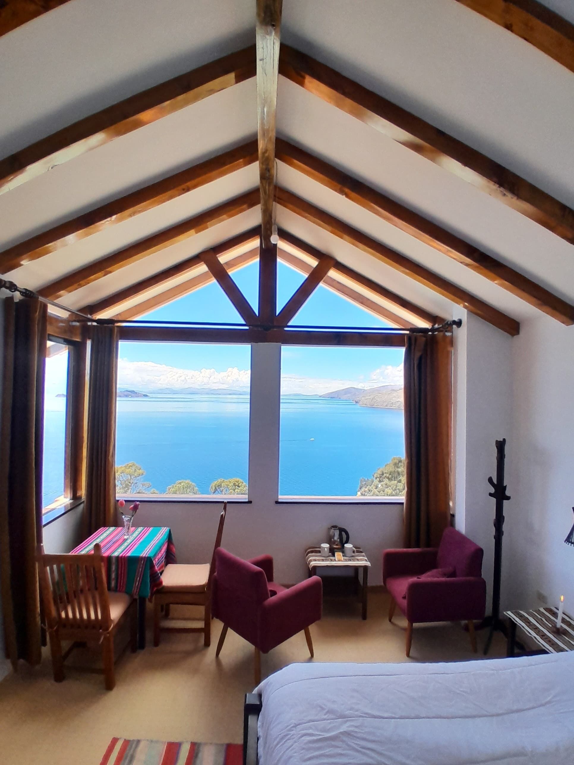 A room with a large window offering a view of a body of water and distant land. There is a bed, a table with a striped tablecloth, two chairs, and a small table with a kettle and cups.