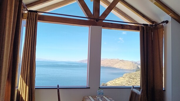 A room with a large window overlooking a body of water and hills, a table with a patterned tablecloth, and two chairs. - Sol y Luna Lodge Isla del Sol Bolivia (Isla del Sol)