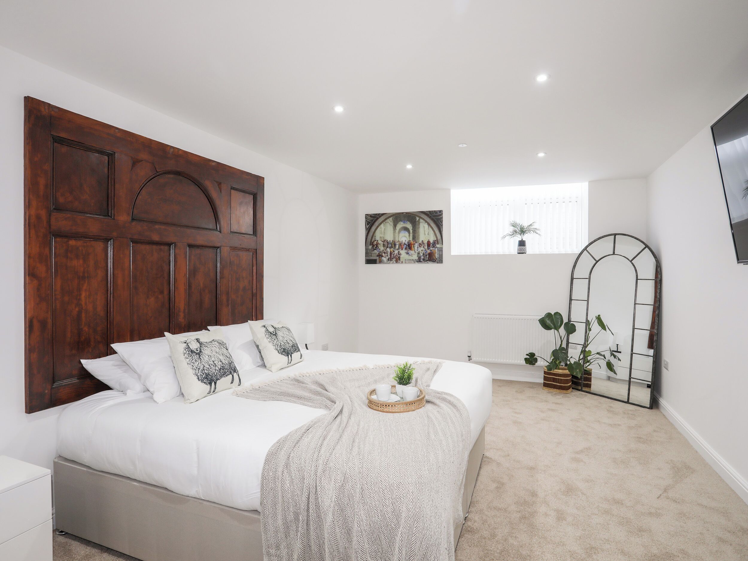 Une chambre avec un grand lit, une tête de lit en bois, un tableau, une fenêtre avec des stores, un miroir et un radiateur.