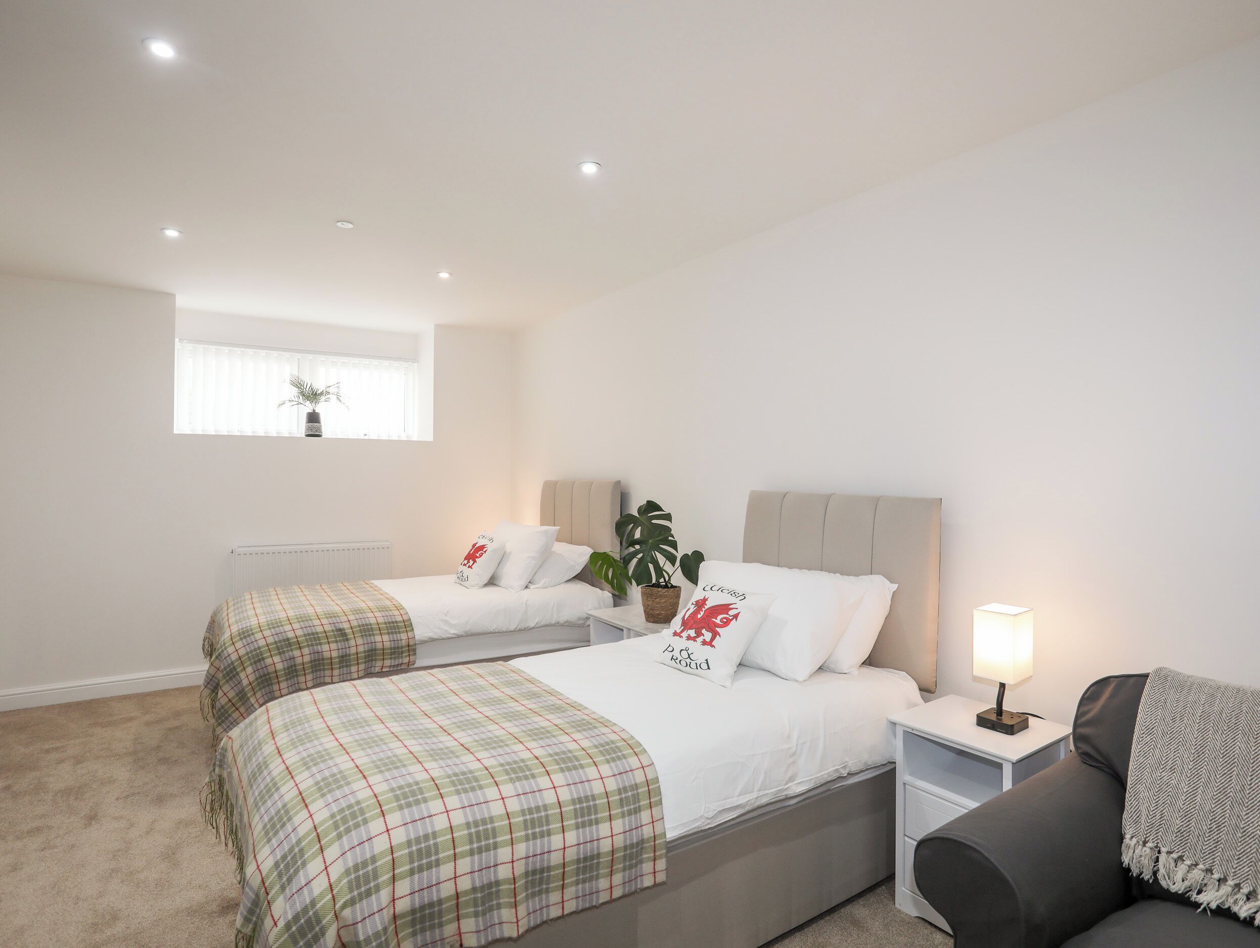 A bedroom with two beds, a window with curtains, a bedside table with a lamp, and a potted plant.