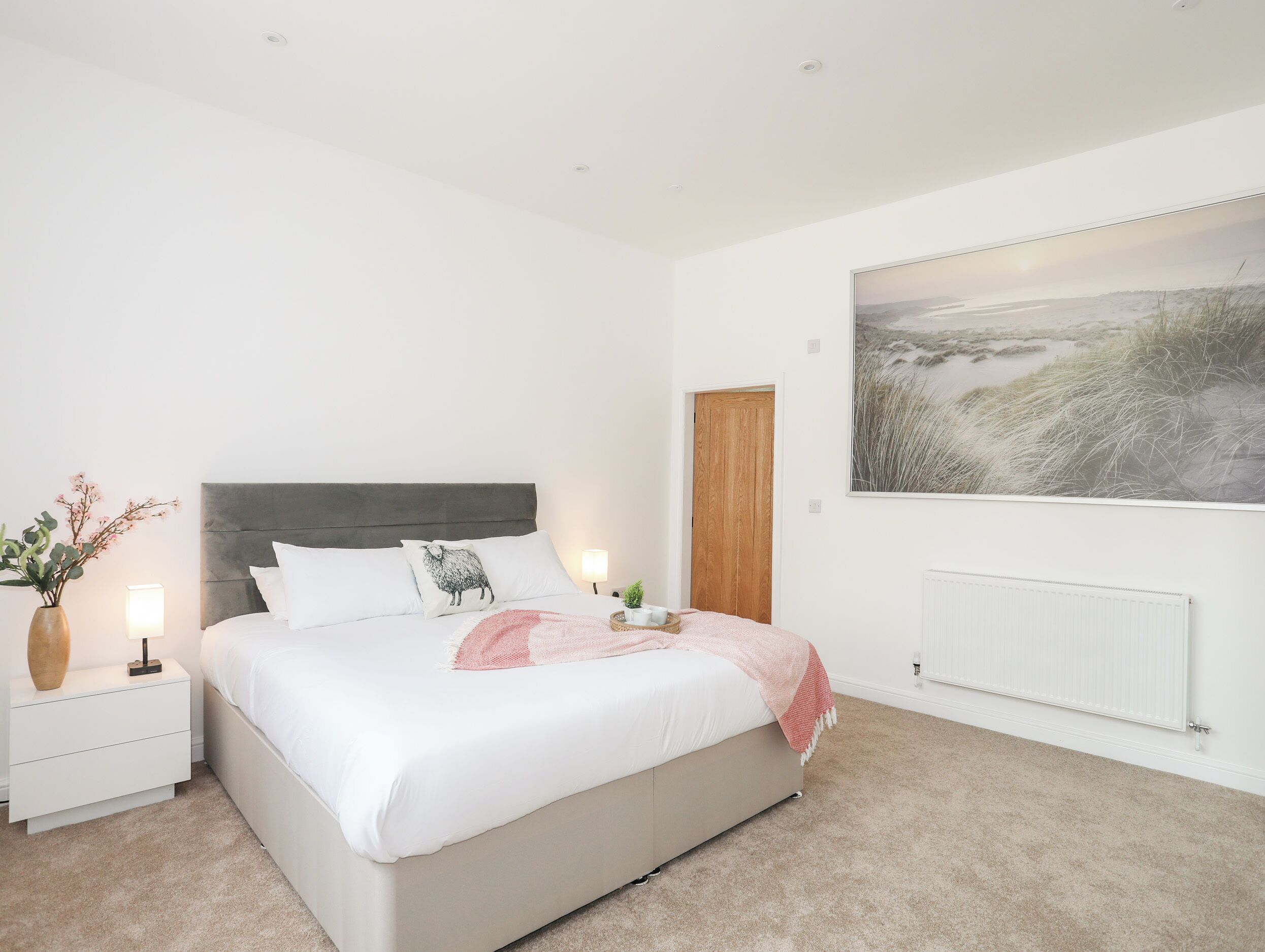 A bedroom with a bed, nightstands, a lamp, a vase with flowers, a painting, and a radiator.