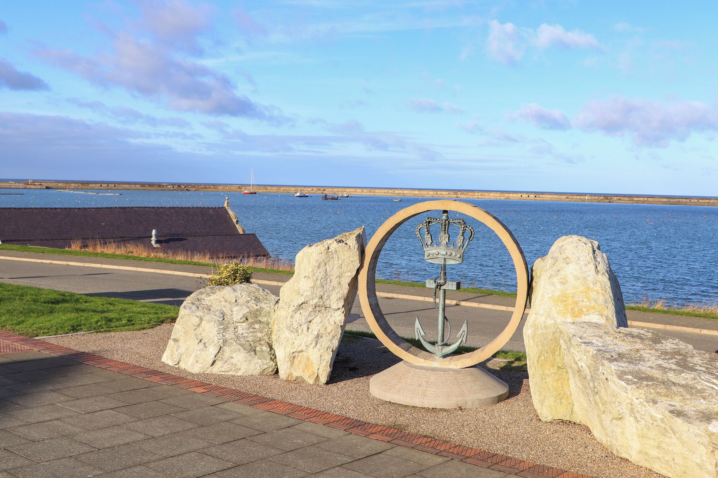 A waterfront view with a circular monument featuring an anchor and crown, surrounded by large rocks and a paved walkway.