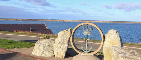 A waterfront view with a circular monument featuring an anchor and crown, surrounded by large rocks and a paved walkway.