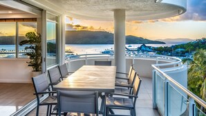 Een balkon met een eettafel en stoelen, met uitzicht op een jachthaven vol boten en een zonsondergang.