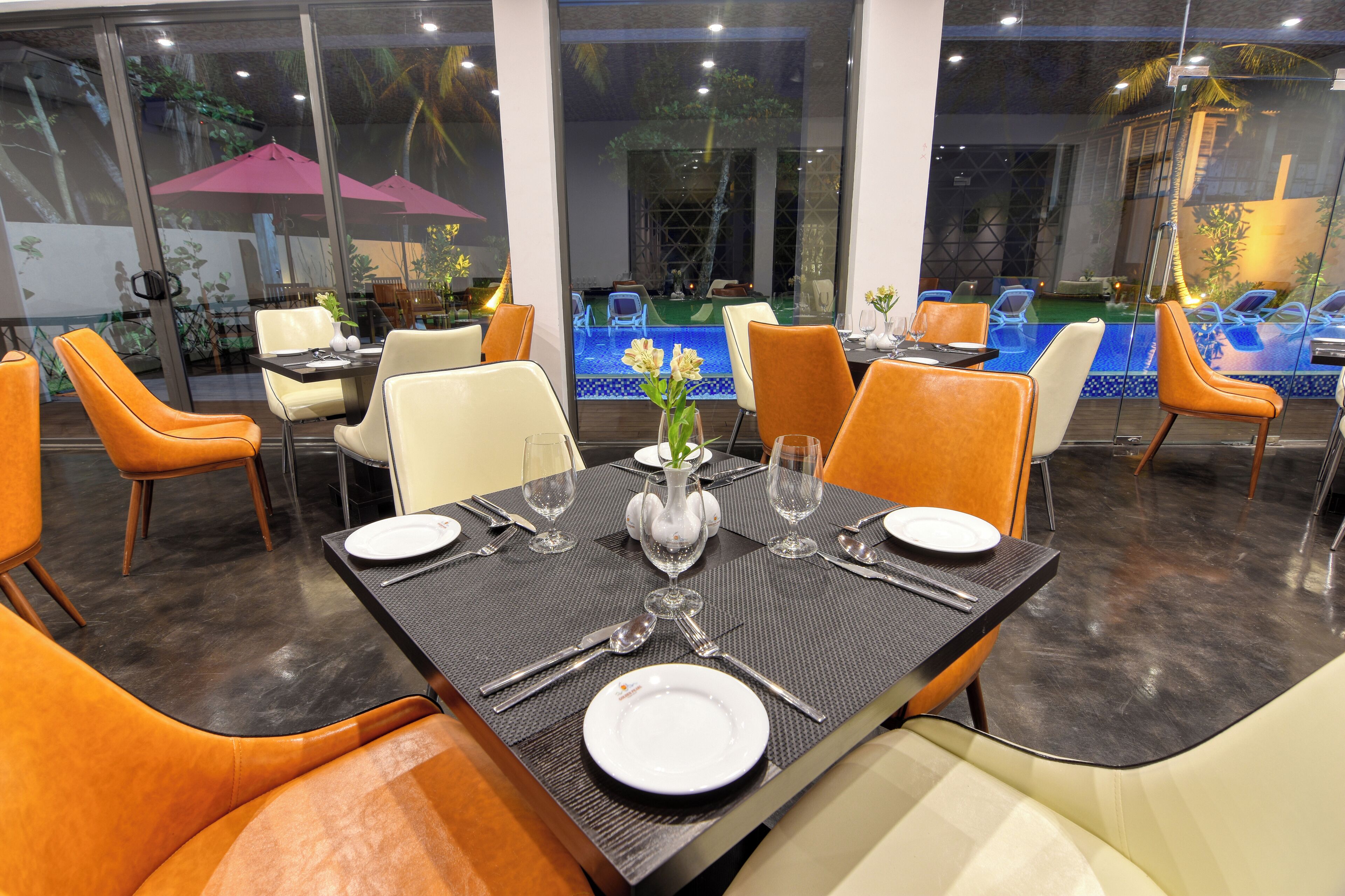 A restaurant with a pool view, tables set with white plates, and orange chairs.