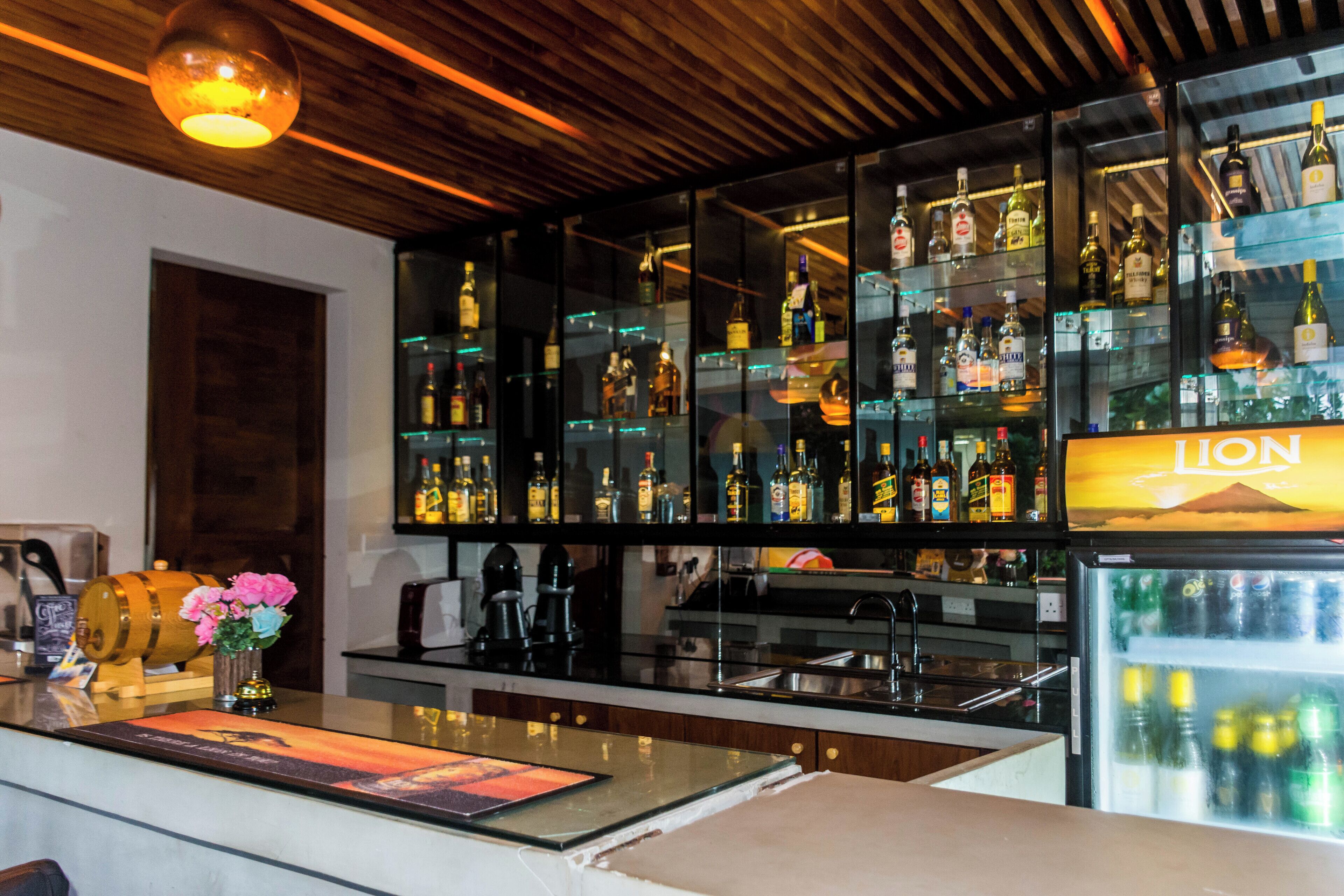 A bar area with a wooden ceiling, a refrigerator with a 'Lion' sign, and various bottles and glasses on shelves.