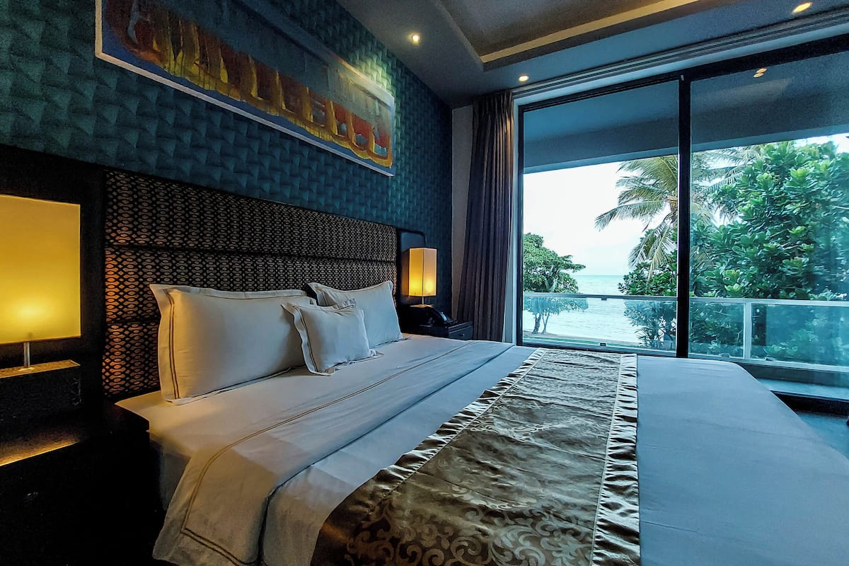 A hotel room with a large bed, a headboard with a textured pattern, a nightstand with a lamp, and a view of the ocean through large windows.