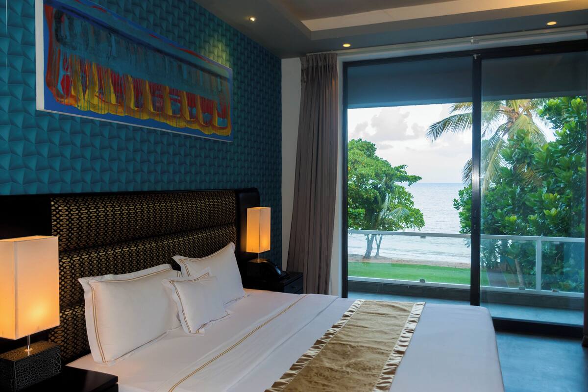 A hotel room with a large bed, a headboard with a textured pattern, a painting on the wall, and a view of the ocean through a large window.